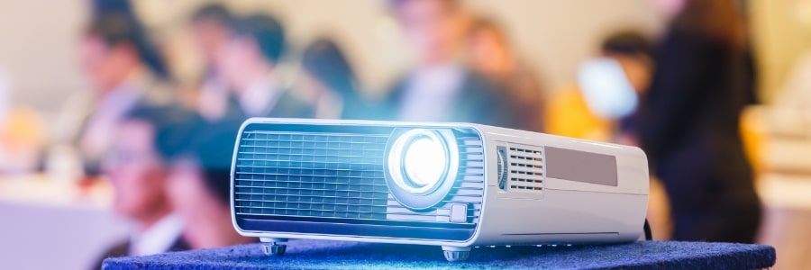 A projector is shining in the foreground with a set of people in formal wear out of focus in the background