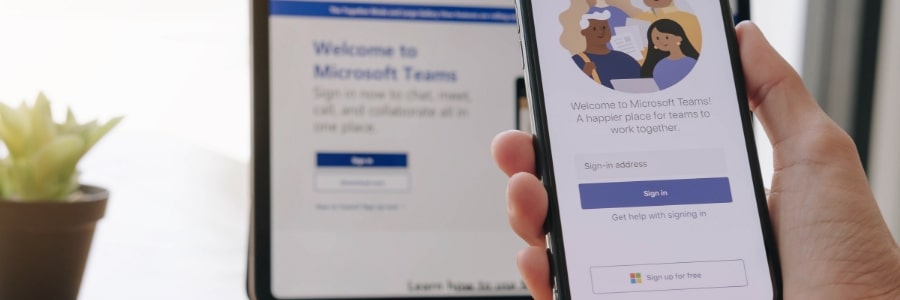 A computer screen with a hand holding a mobile phone in front of it. Both screens have Microsoft Teams showing