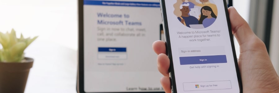 A computer screen with a hand holding a mobile phone in front of it. Both screens have Microsoft Teams showing